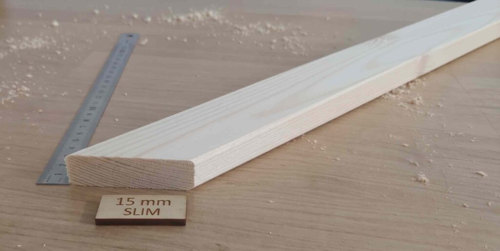 A close-up photograph of a light-colored pine wood stretcher bar with a mitered corner on a workshop table surface. In the foreground, a small engraved wooden tag reads '15 mm SLIM'. A metal ruler and scattered sawdust are visible in the background.