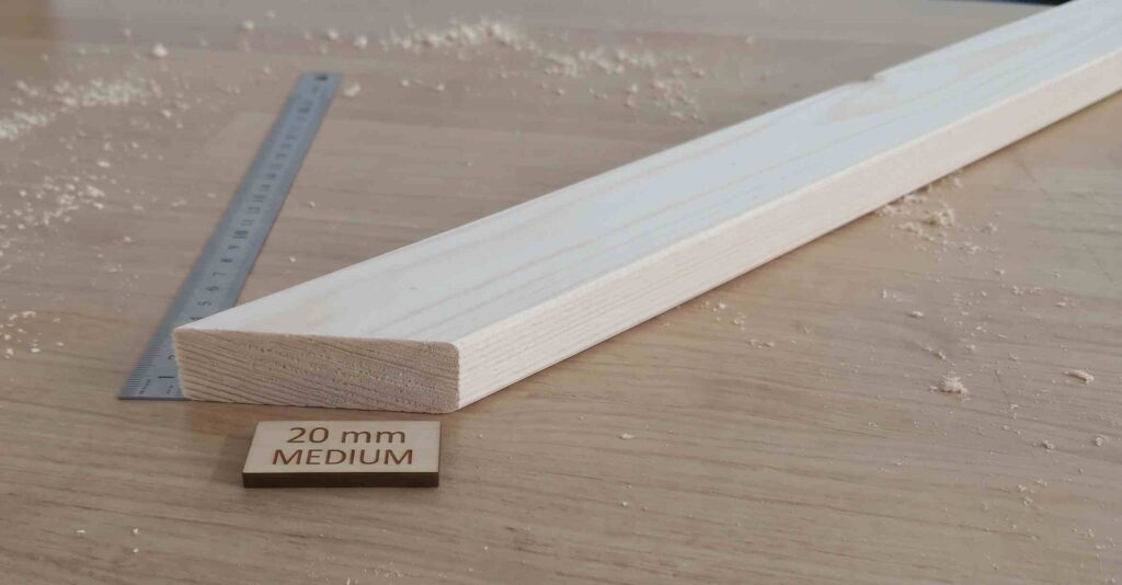 A single piece of light-colored wood, cut at an angle, representing a 20mm medium stretcher bar profile. It rests on a wooden surface with wood shavings, placed next to a metal measuring ruler and a small wooden tag that reads "20 mm MEDIUM".