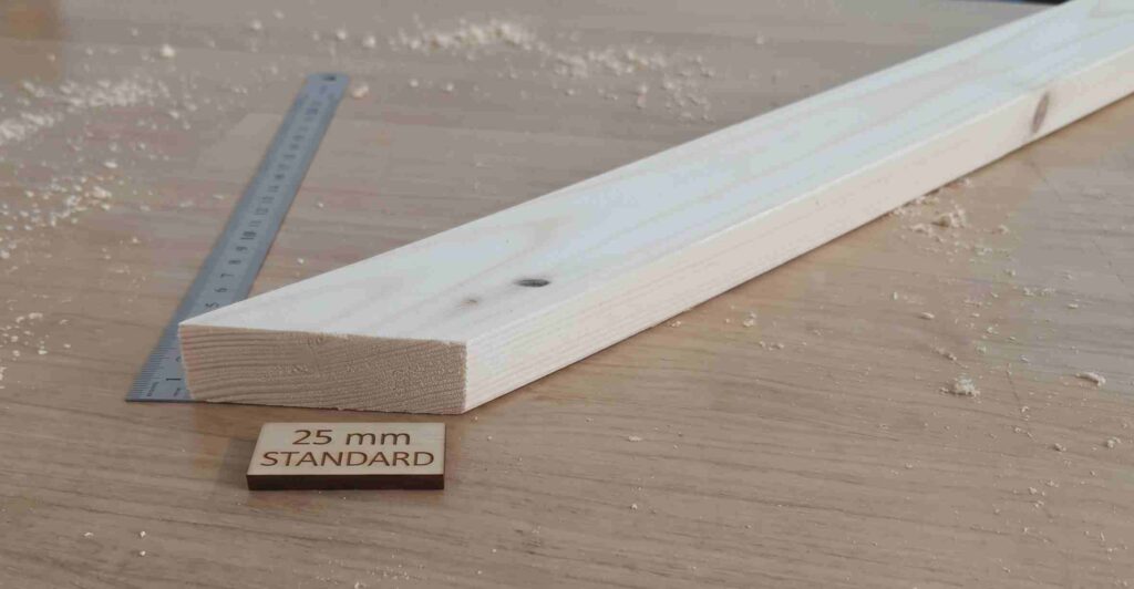 A 25 mm thick wooden stretcher bar resting on a wooden surface surrounded by sawdust, accompanied by a metal ruler and a small wooden tag that reads "25 mm STANDARD".