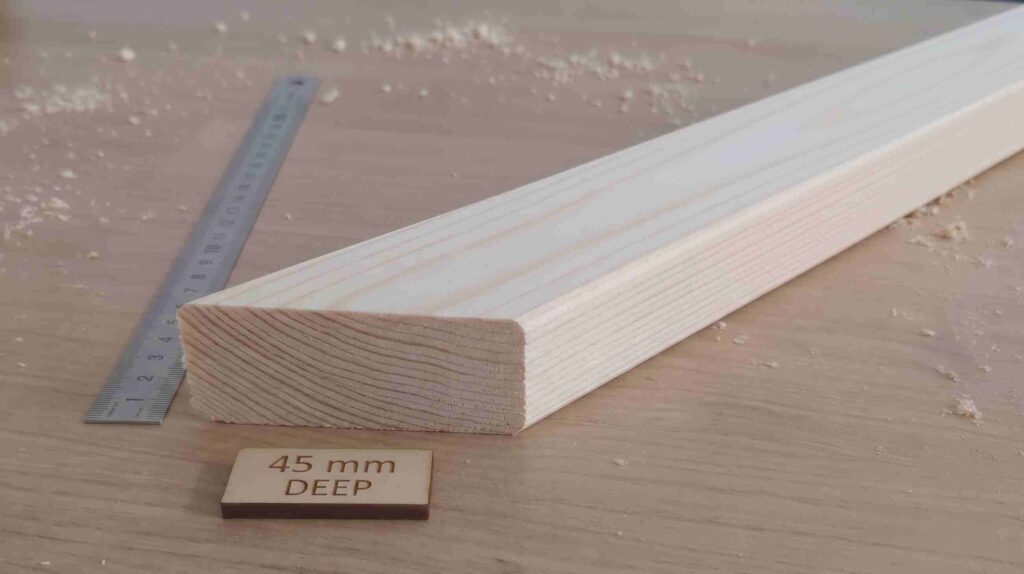 A close-up cross-section view of a heavy-duty unfinished pine wood stretcher bar. The end-grain is visible with prominent natural growth rings. It is next to a metal ruler and a small wooden tag that reads "45 mm DEEP". Wood shavings are on the wooden workbench.