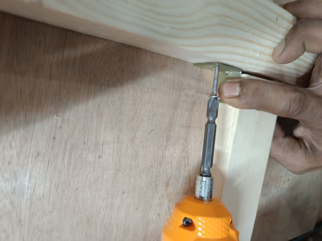 an orange and black power drill to screw a gold-colored L-bracket onto the inside corner joint of a light-wood frame, which is resting on a plywood background.